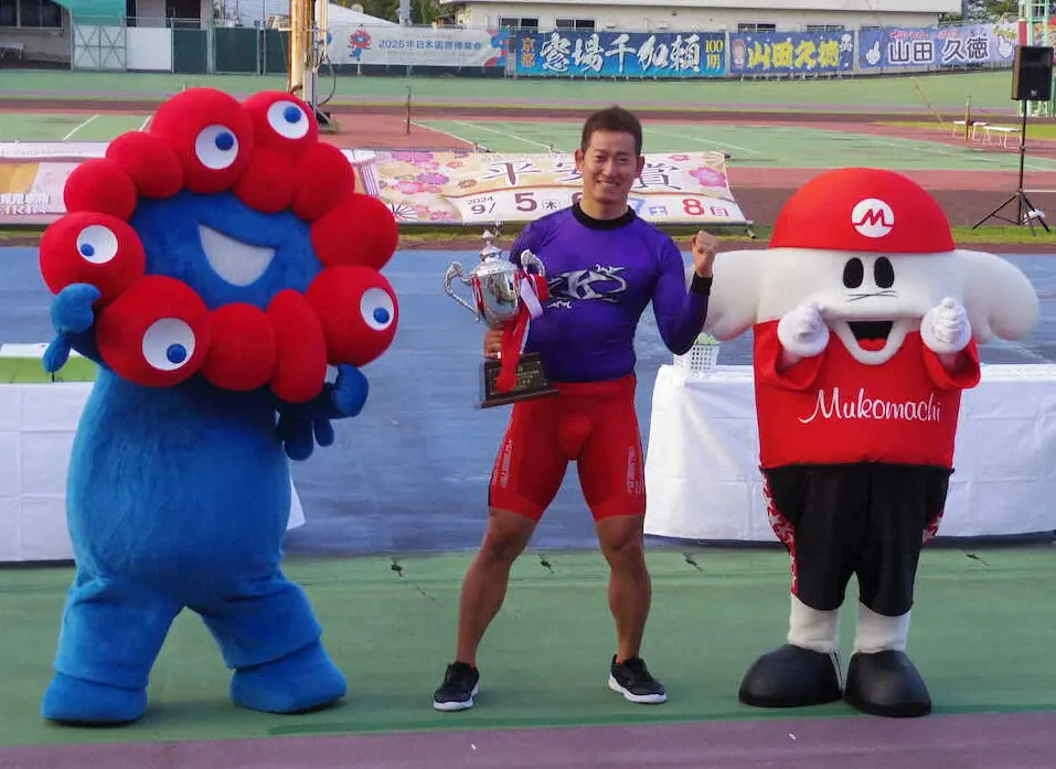大阪・関西万博公式キャラクター「ミャクミャク」（左）と向日町競輪マスコットキャラクター「ムコリン・ムッチー」（右）の間でガッツポーズをする脇本